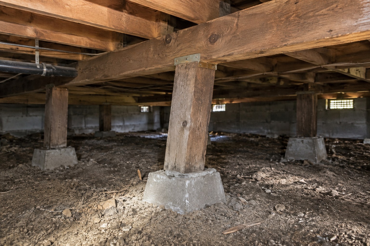 Crawlspace of a raised-floor house with wood posts on concrete piers under a beam, one post leaning